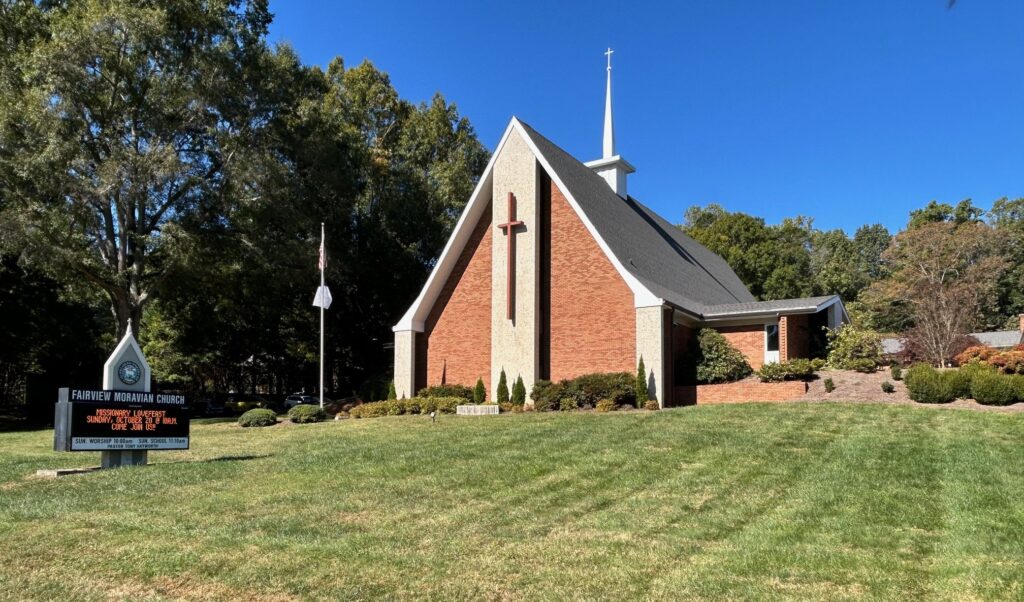 Photo of a brick church with a steeple and a big wooden cross on the front. There is green grass and shrubs around the building and trees behind it. An American flag flies on a pole in front of the building. The church sign is also pictured, the text is too small to read in full but the top says Fairview Moravian Church. The sky is blue and cloudless.
